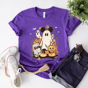 May include: Purple t-shirt with a graphic of a ghost Mickey Mouse holding a jack-o-lantern. The ghost is wearing a black hat with a leaf on top. The graphic also includes a tombstone that says "RIP" and a few scattered leaves.
