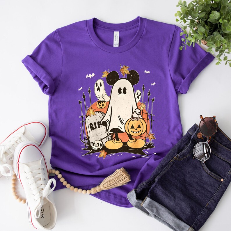 May include: Purple t-shirt with a graphic of a ghost Mickey Mouse holding a jack-o-lantern. The ghost is wearing a black hat with a leaf on top. The graphic also includes a tombstone that says "RIP" and a few scattered leaves.