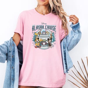 Pu&ograve; includere: T-shirt rosa chiaro con la scritta "Disney Alaska Cruise" e una grafica di Topolino, Minnie e altri personaggi Disney. L'anno "2025" &egrave; anch'esso visualizzato sulla maglietta.