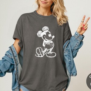 May include: Dark gray t-shirt featuring a white sketch of Mickey Mouse. The shirt has short sleeves and a relaxed fit. The image also shows a denim jacket. The text on the image says "Comfort Colors Pepper".