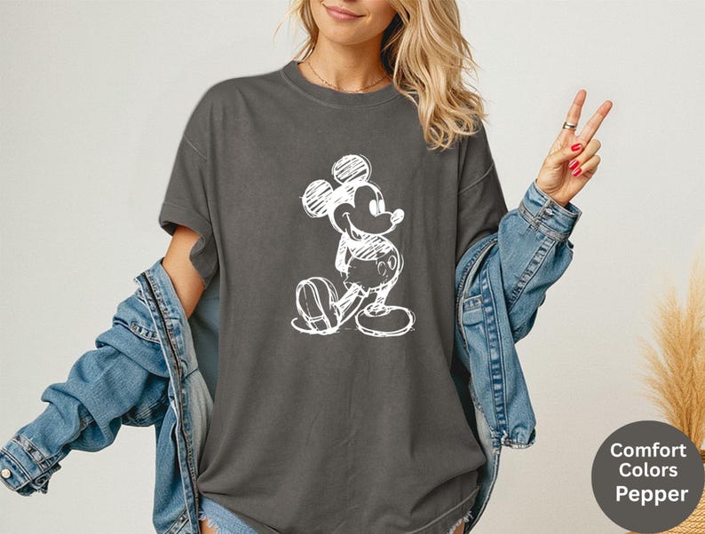 May include: Dark gray t-shirt featuring a white sketch of Mickey Mouse. The shirt has short sleeves and a relaxed fit. The image also shows a denim jacket. The text on the image says "Comfort Colors Pepper".