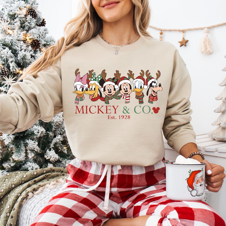 K&ouml;nnte beinhalten: Beiges Sweatshirt mit einem festlichen Disney-Motiv von Charakteren mit Rentiergeweihen und Schals. Der Text "MICKEY & CO." und "Est. 1928" sind darunter aufgedruckt. Dazu eine rot-wei&szlig; karierte Pyjamahose.