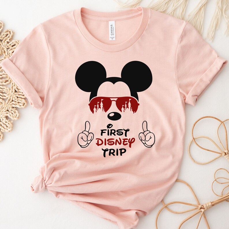 Op de afbeelding: Roze T-shirt met een zwarte Mickey Mouse-afbeelding die een zonnebril draagt met een rood kasteel silhouet. De tekst "First Disney Trip" staat onder de afbeelding.
