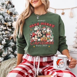 Puede incluir: Sudadera verde con Mickey Mouse y sus amigos y el texto "mickey's VERY MERRY Christmas Party 2025". El dise&ntilde;o incluye a Mickey, Minnie, Donald, Goofy y Pluto. Una taza blanca con un dibujo de Pap&aacute; Noel se sostiene en la mano.