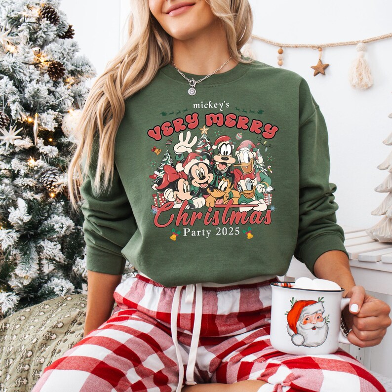 Puede incluir: Sudadera verde con Mickey Mouse y sus amigos y el texto "mickey's VERY MERRY Christmas Party 2025". El dise&ntilde;o incluye a Mickey, Minnie, Donald, Goofy y Pluto. Una taza blanca con un dibujo de Pap&aacute; Noel se sostiene en la mano.