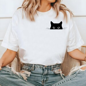 Pu&ograve; includere: Maglietta bianca con un gatto nero che spunta dalla tasca. Il gatto ha occhi grandi e baffi.