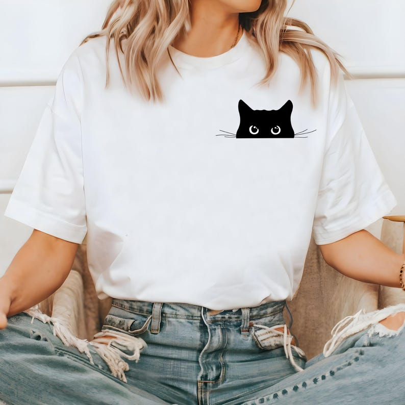 Pu&ograve; includere: Maglietta bianca con un gatto nero che spunta dalla tasca. Il gatto ha occhi grandi e baffi.