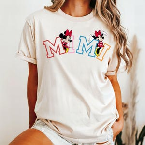 May include: A cream colored t-shirt with a graphic of two Mickey Mouse characters in front of the word "MIMI" in colorful letters.
