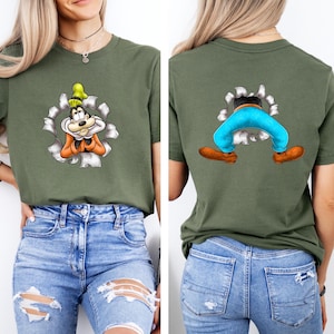 K&ouml;nnte beinhalten: Olivgr&uuml;nes T-Shirt mit einem Cartoon-Charakter-Design. Die Vorderseite zeigt die Figur, die durch ein zerrissenes Loch blickt, und die R&uuml;ckseite zeigt die Beine und F&uuml;&szlig;e der Figur. Das Design enth&auml;lt orange, gelbe, blaue und braune Akzente.