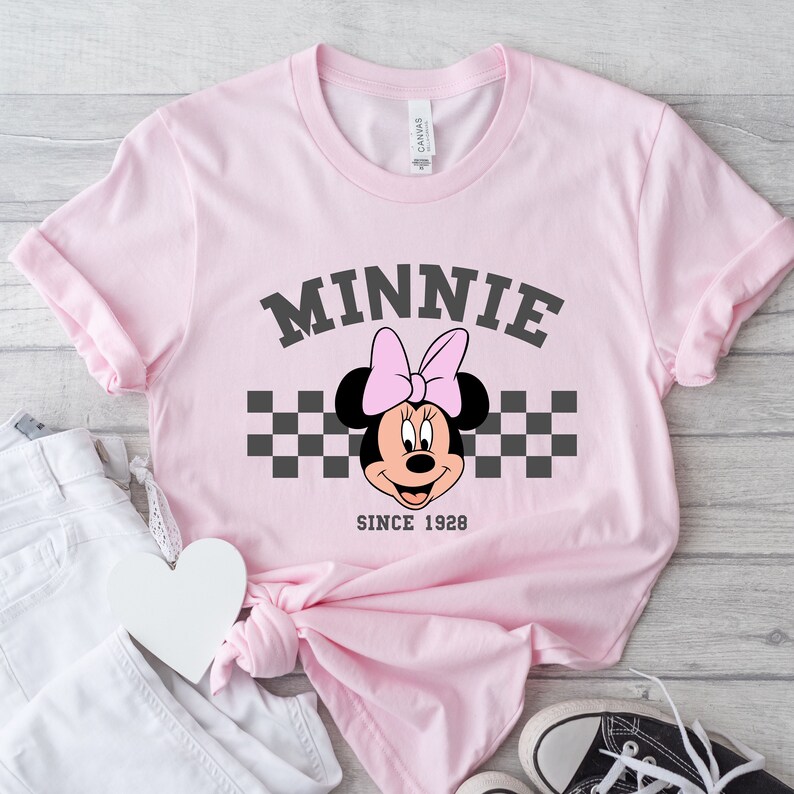 May include: A light pink t-shirt featuring a Minnie Mouse graphic. The design includes the name "MINNIE" in gray block letters above Minnie's face, a pink bow, and a checkered pattern. The text "SINCE 1928" is below the character.