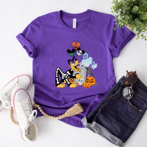 May include: A purple t-shirt featuring a Halloween-themed graphic. The design includes a cartoon dog in a skeleton costume, pumpkins, a tombstone, and balloons with character faces. The shirt is paired with denim shorts and white sneakers.