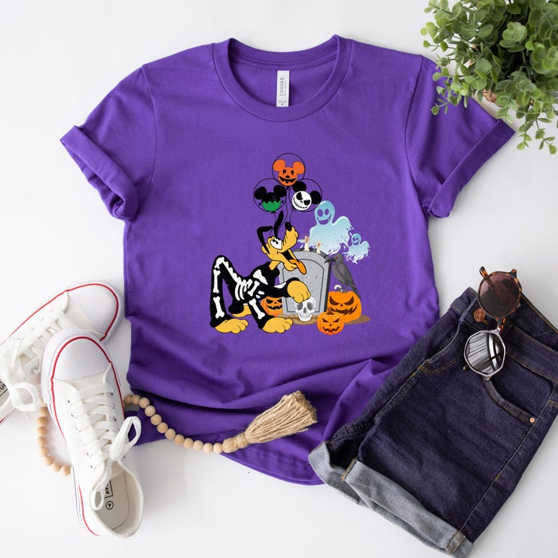May include: A purple t-shirt featuring a Halloween-themed graphic. The design includes a cartoon dog in a skeleton costume, pumpkins, a tombstone, and balloons with character faces. The shirt is paired with denim shorts and white sneakers.