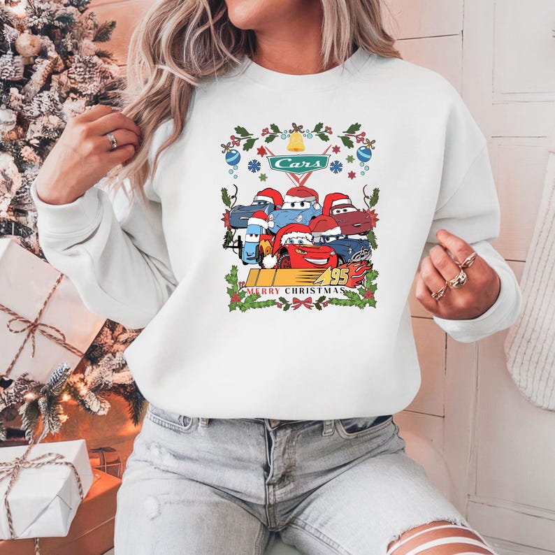 May include: White sweatshirt featuring a graphic of Cars characters wearing Santa hats, with the text "Cars" and "Merry Christmas 95". The design includes festive holly and ornaments.