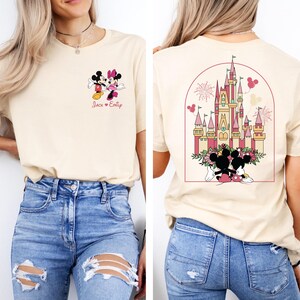 K&ouml;nnte beinhalten: Ein hellbeiges T-Shirt mit einem Motiv von Mickey und Minnie Mouse auf der Vorderseite und einem Motiv von Schloss Aschenputtel mit Mickey und Minnie Mouse auf der R&uuml;ckseite. Der Text "Jack & Emily" ist unter dem Mickey- und Minnie-Mouse-Motiv auf der Vorderseite des Shirts gedruckt.