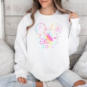 May include: White sweatshirt featuring a colorful Mickey Mouse head design with a castle silhouette and the text "Disney 2026". The design incorporates pastel colors and sparkles, creating a whimsical and celebratory aesthetic.