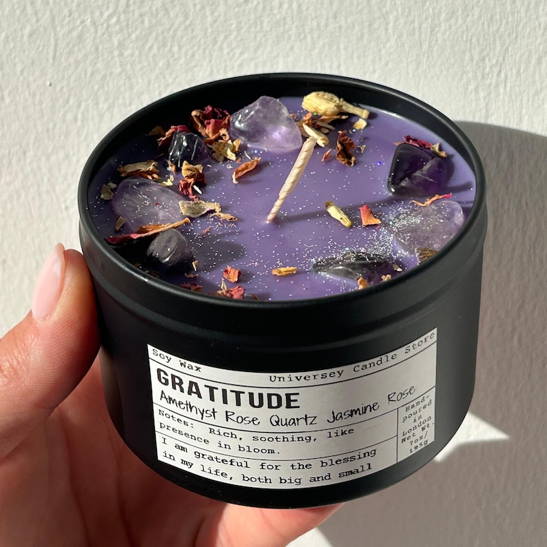 May include: A black metal tin candle with purple wax, topped with amethyst crystals, dried rose petals, and a single wick. The white label reads: Soy Wax, Universey Candle Store, GRATITUDE, Amethyst Rose Quartz Jasmine Rose, Notes: Rich, soothing, like presence in bloom. I am grateful for the blessing in my life, both big and small. Hand-poured in London, Net Wt. 7oz.