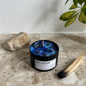 May include: A black metal candle tin filled with blue wax, dark blue swirls, and embedded dark blue stones. The white label reads Inner Wisdom, Lapis Lazuli Cornflower, and includes text about clarity. A single white wick is centered in the wax.