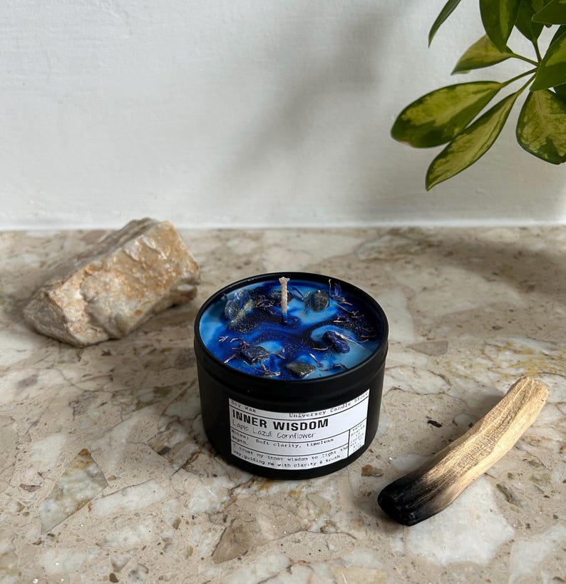 May include: A black metal candle tin filled with blue wax, dark blue swirls, and embedded dark blue stones. The white label reads Inner Wisdom, Lapis Lazuli Cornflower, and includes text about clarity. A single white wick is centered in the wax.
