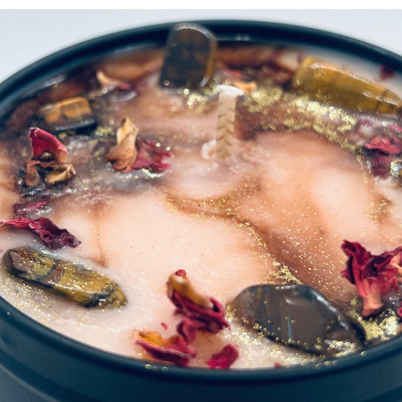 May include: Close-up of a pink candle with gold flecks, brown and gold stones, and dried rose petals. The candle is in a black jar.