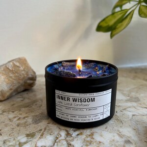 May include: A black metal tin candle with a lit wick and blue wax topped with dark blue crystals. The white label reads: Soy Wax, Universey Candle Store, INNER WISDOM, Lapis Lazuli Cornflower, Notes: Soft clarity, timeless depth, I trust my inner wisdom to light the way, guiding me with clarity & truth.