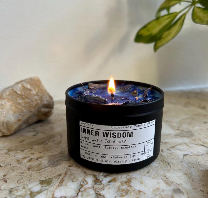 May include: A black metal tin candle with a lit wick and blue wax topped with dark blue crystals. The white label reads: Soy Wax, Universey Candle Store, INNER WISDOM, Lapis Lazuli Cornflower, Notes: Soft clarity, timeless depth, I trust my inner wisdom to light the way, guiding me with clarity & truth.