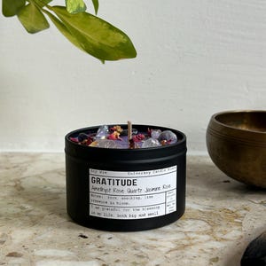 May include: A black metal tin candle filled with purple soy wax, topped with amethyst and rose quartz crystals and dried flower petals. The label reads "GRATITUDE Amethyst Rose Quartz Jasmine Rose" and "net wt 135g". A single cotton wick is centered.