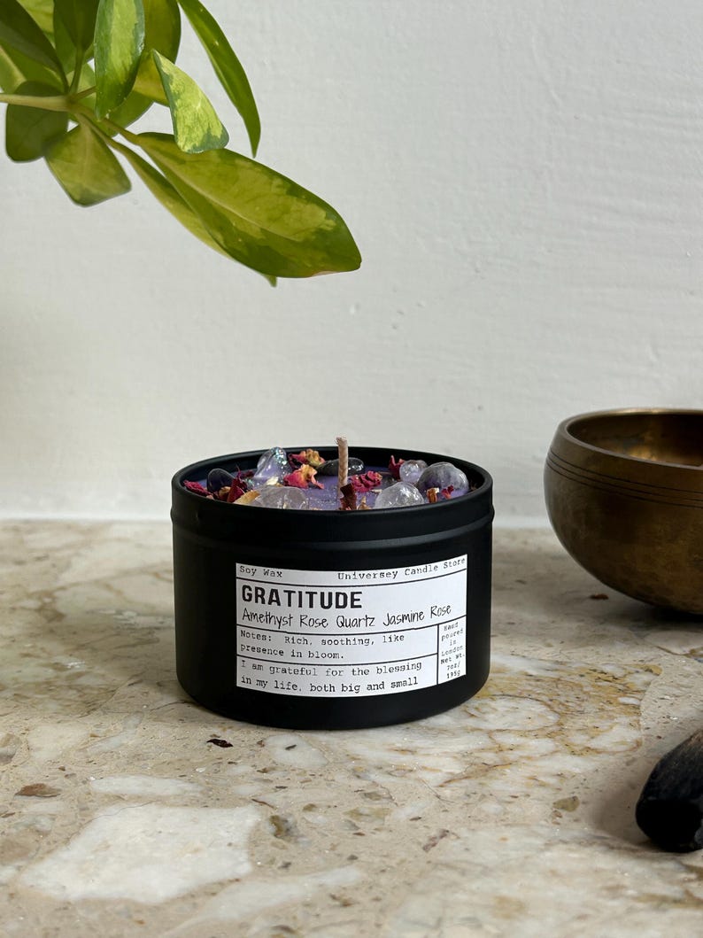 May include: A black metal tin candle filled with purple soy wax, topped with amethyst and rose quartz crystals and dried flower petals. The label reads "GRATITUDE Amethyst Rose Quartz Jasmine Rose" and "net wt 135g". A single cotton wick is centered.
