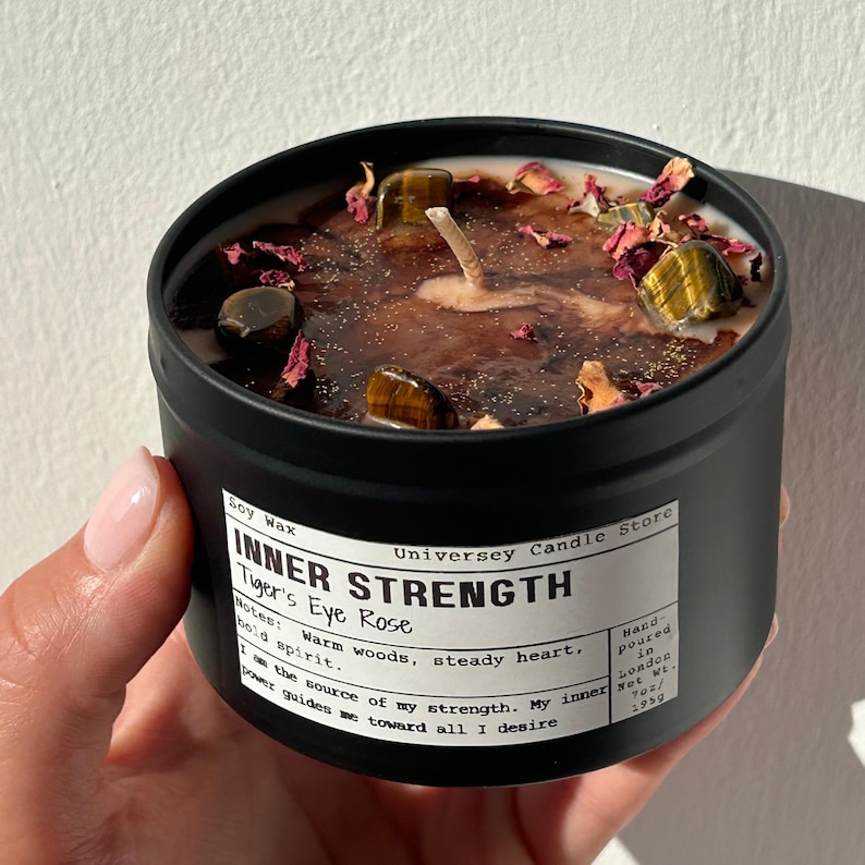 May include: A black metal tin candle filled with soy wax, topped with tiger's eye stones and dried rose petals. The label reads: INNER STRENGTH, Tiger's Eye Rose, Notes: Warm woods, steady heart, bold spirit. I am the source of my strength. Net Wt. 7oz.