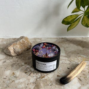 May include: A black metal candle tin filled with purple wax, topped with small clear crystals and dried flower petals. The white label features the text GRATITUDE, Amethyst Rose Quartz Jasmine Rose, and additional descriptive text.