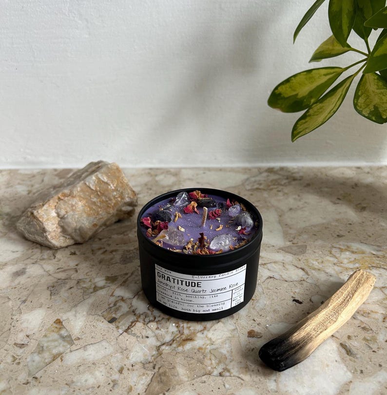 May include: A black metal candle tin filled with purple wax, topped with small clear crystals and dried flower petals. The white label features the text GRATITUDE, Amethyst Rose Quartz Jasmine Rose, and additional descriptive text.