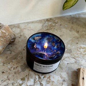 May include: A round black candle tin containing blue wax with embedded dark stones and a lit wick. The white label on the side features the text Inner Wisdom, Lapis Lazuli Cornflower, and additional printed details in black ink.