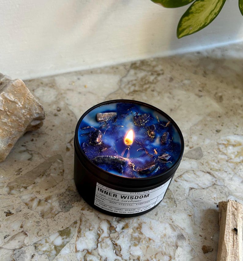 May include: A round black candle tin containing blue wax with embedded dark stones and a lit wick. The white label on the side features the text Inner Wisdom, Lapis Lazuli Cornflower, and additional printed details in black ink.