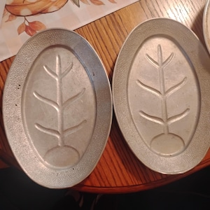 May include: Two oval-shaped silver-toned metal platters featuring a textured rim and a central embossed fish skeleton design. The platters have a smooth, recessed center area and a raised, pebbled border around the outer edge.