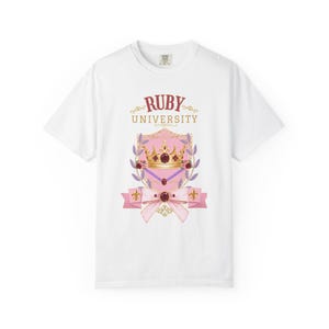 May include: A white short-sleeve t-shirt featuring a graphic print of a gold crown centered on a pink shield. The text RUBY UNIVERSITY appears above the shield, which is decorated with floral accents, a pink ribbon, and a central red gemstone.