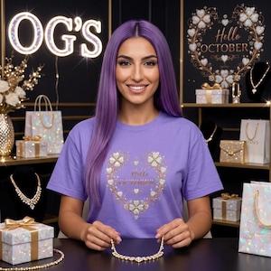 May include: A light purple t-shirt featuring a gold heart-shaped wreath graphic made of small flowers and leaves, with the text Hello OCTOBER centered inside. The design includes sparkling accents throughout the floral heart motif.
