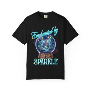 May include: A black short-sleeve t-shirt featuring a graphic of a crystal ball containing a castle, trees, and a river under a crescent moon. The text "Enchanted by" appears in light blue above the ball, and "SPARKLE" is printed below in blue.