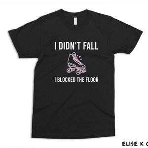 K&ouml;nnte beinhalten: Schwarzes T-Shirt mit wei&szlig;em Text, der "Ich bin nicht gefallen, ich habe den Boden blockiert" lautet. Eine rosa Rollschuhgrafik befindet sich &uuml;ber dem Text.