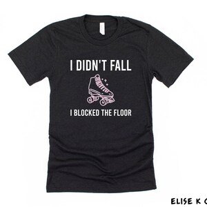 K&ouml;nnte beinhalten: Schwarzes T-Shirt mit dem Text "Ich bin nicht gefallen, ich habe den Boden blockiert" in Wei&szlig;. Eine rosa Rollschuhgrafik befindet sich &uuml;ber dem Text.