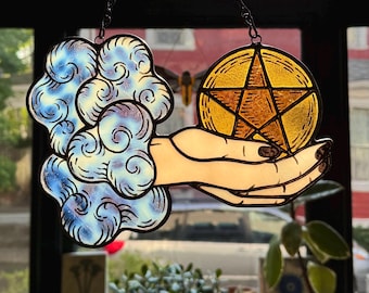 May include: Stained glass suncatcher featuring a hand holding a golden pentagram and a blue and white cloud. The artwork is outlined in black and hangs from a chain. The background shows a window with plants.