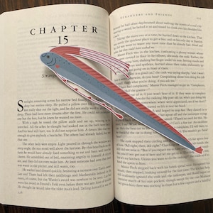 May include: A whimsical fish-shaped bookmark in shades of gray, red, and blue, resting on an open book. The bookmark's design includes a fish head and tail, with a streamlined body. The book is open to Chapter 15.