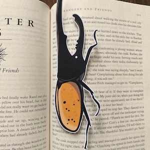 May include: Black beetle-shaped bookmark with an orange and black body, and long, curved antennae. The bookmark is placed on the pages of a book. The bookmark is a unique accessory for book lovers.