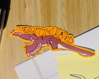 May include: A sticker with the word "OVERSTIMULATED" in orange, featuring a cartoon dinosaur in shades of purple and white. The sticker is on a wooden surface with a pen and paper.