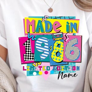 May include: White t-shirt with a colorful graphic design. The design features the words "MADE IN 1986" in bold, retro-style lettering, along with the words "LIMITED EDITION" and a space for a name. The numbers are in vibrant colors.