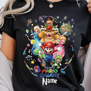 May include: Black t-shirt featuring a colorful graphic of Super Mario characters, including Mario, Princess Peach, and Bowser. The text "THE SUPER MARIO GALAXY Name" is printed below the characters. The design is surrounded by a rainbow-colored border.