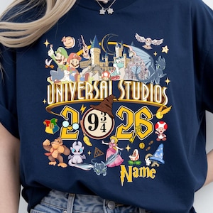 Może przedstawiać: Granatowa koszulka z kolorową grafiką. Projekt zawiera napis "Universal Studios 2026", postacie z kreskówek, pociąg Hogwart Express, tiarę przydziału i numer 9 3/4.