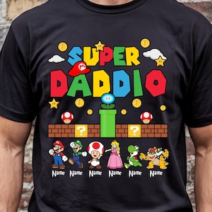 May include: Black t-shirt featuring a colorful graphic with the words "SUPER DADDIO" and various cartoon characters. The design includes stars, coins, and a brick platform. Below the characters are spaces for names.