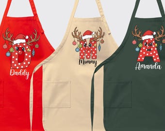 Puede incluir: Tres delantales navideños en rojo, beige y verde oscuro. Cada delantal presenta una letra con cara de reno, un gorro de Papá Noel y los nombres "Daddy", "Mommy" y "Amanda". Los delantales tienen bolsillos y correas ajustables para el cuello.