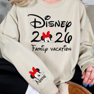 May include: Beige sweatshirt with black Disney logo, "2026", and "Family Vacation" text. Features a Minnie Mouse graphic with a red bow. The sleeve has a Minnie Mouse graphic and the word "Name".