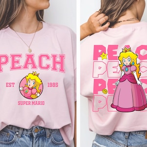 May include: Light pink t-shirt featuring the word "PEACH" in large pink block letters. The front of the shirt has a circular graphic of Princess Peach from Super Mario. The back of the shirt has a larger graphic of Princess Peach.