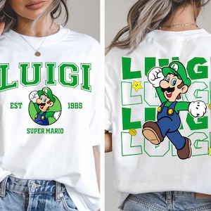 May include: White t-shirt featuring a green "LUIGI" graphic with a cartoon image of Luigi and the text "EST 1985 SUPER MARIO" on the front. The back of the shirt has a green Luigi graphic and the word "LUIGI" repeated.